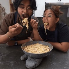 Two people with glasses are eating spaghetti from a large bowl in a kitchen