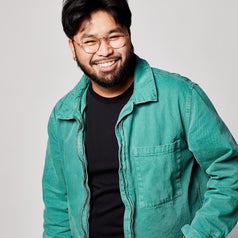 Gideon with glasses and a beard wears a casual outfit including a green jacket and a black shirt