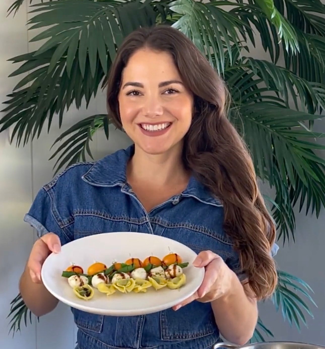 A smiling woman in a denim jumpsuit holds a plate of freshly prepared appetizers, with a bowl of tomatoes and a colander of vegetables on the table