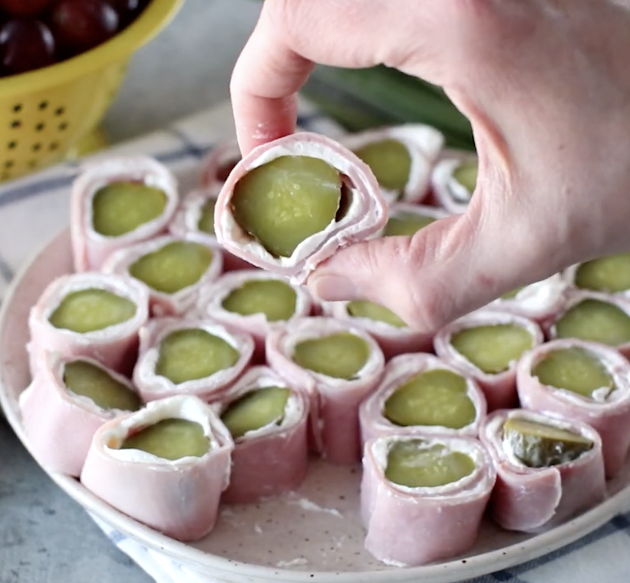 A hand holds a ham and pickle roll-up with cream cheese, while similar roll-ups are arranged on a plate in the background next to a yellow colander of grapes