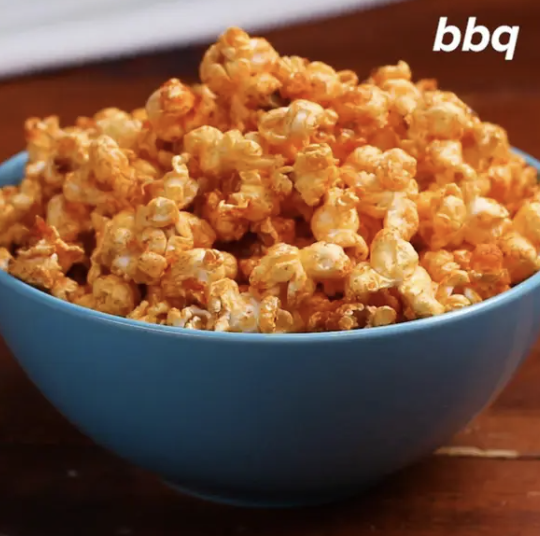 A bowl of BBQ-flavored popcorn sits on a wooden surface, with &quot;bbq&quot; written in white text in the upper right corner