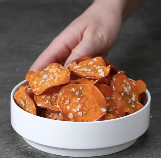 A hand reaching for a bowl of sweet potato chips topped with sea salt