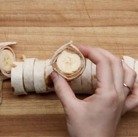 A hand holding a pinwheel snack made of tortilla wrapped around banana slices, with more pinwheel snacks placed on a wooden cutting board