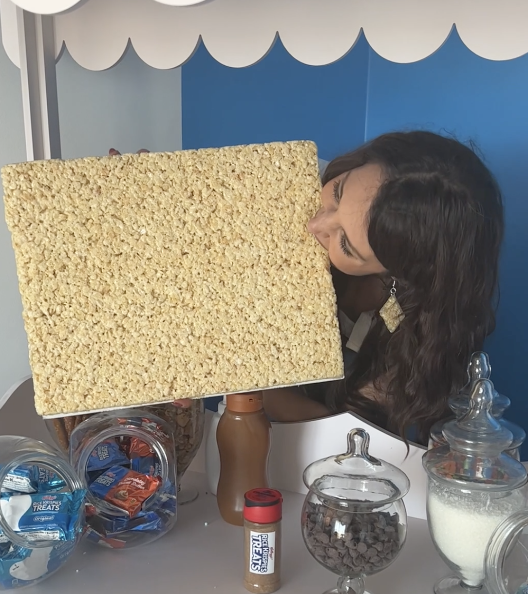 Woman holding and taking a bite from a giant Rice Krispies Treat. Jars of candy and cereal are on the table in front of her