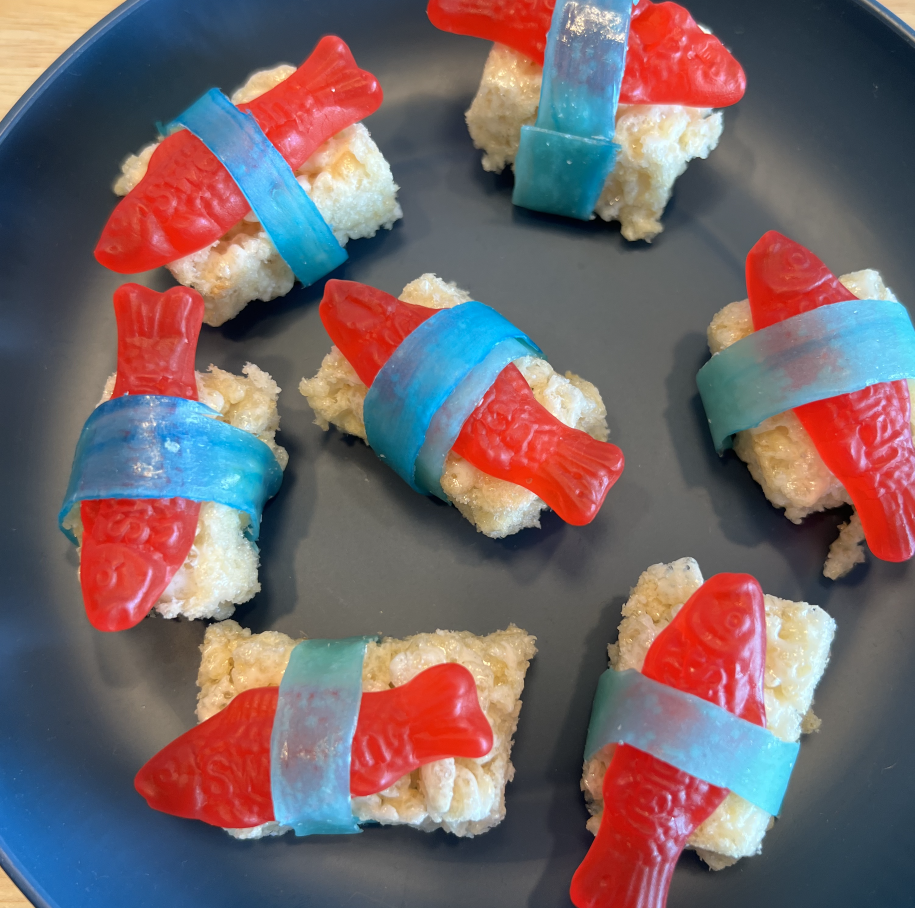 Rice Krispie sushi pieces topped with red gummy fish and wrapped in blue fruit roll-up strips on a black plate