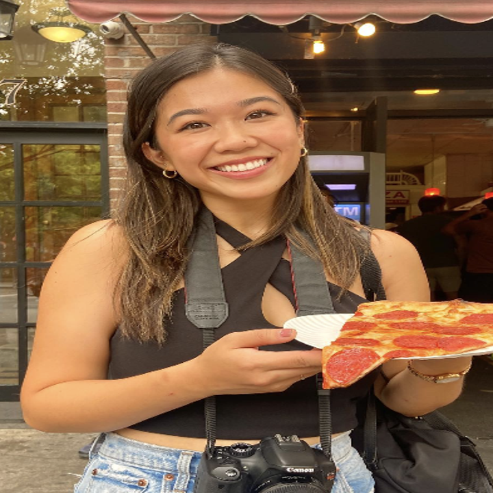 Woman standing outside a building, smiling, holding a slice of pizza on a paper plate. She is wearing a black top, denim shorts, and has cameras hanging around her neck